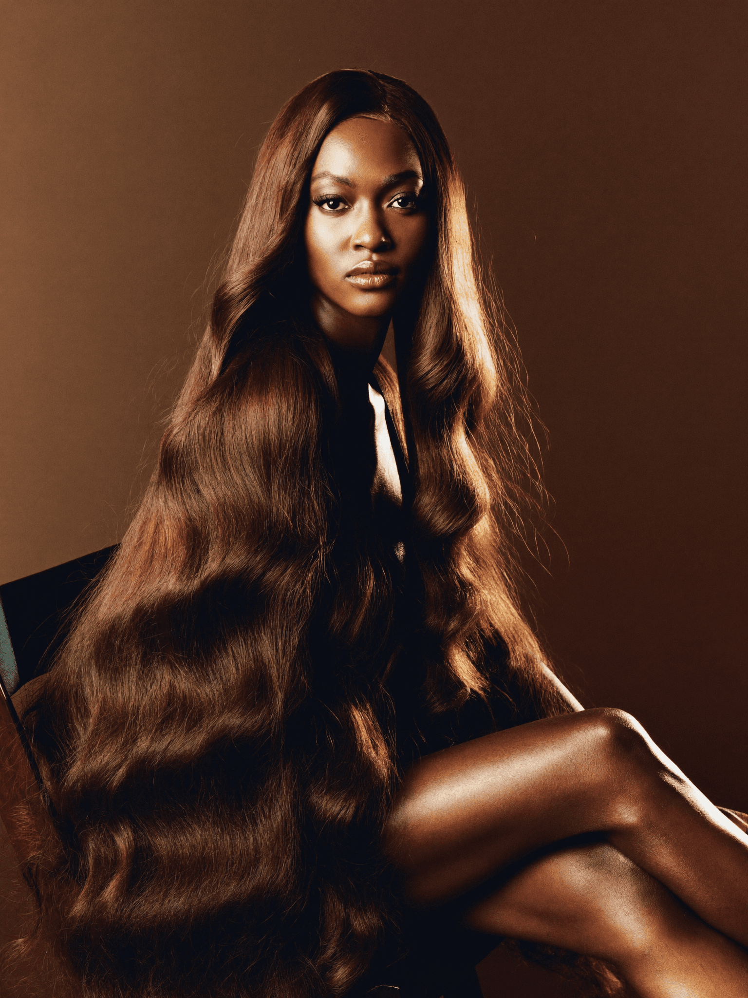 A woman seated in a studio portrait wearing a long, voluminous Naronai wig in rich warm brown tones, her gaze direct and commanding